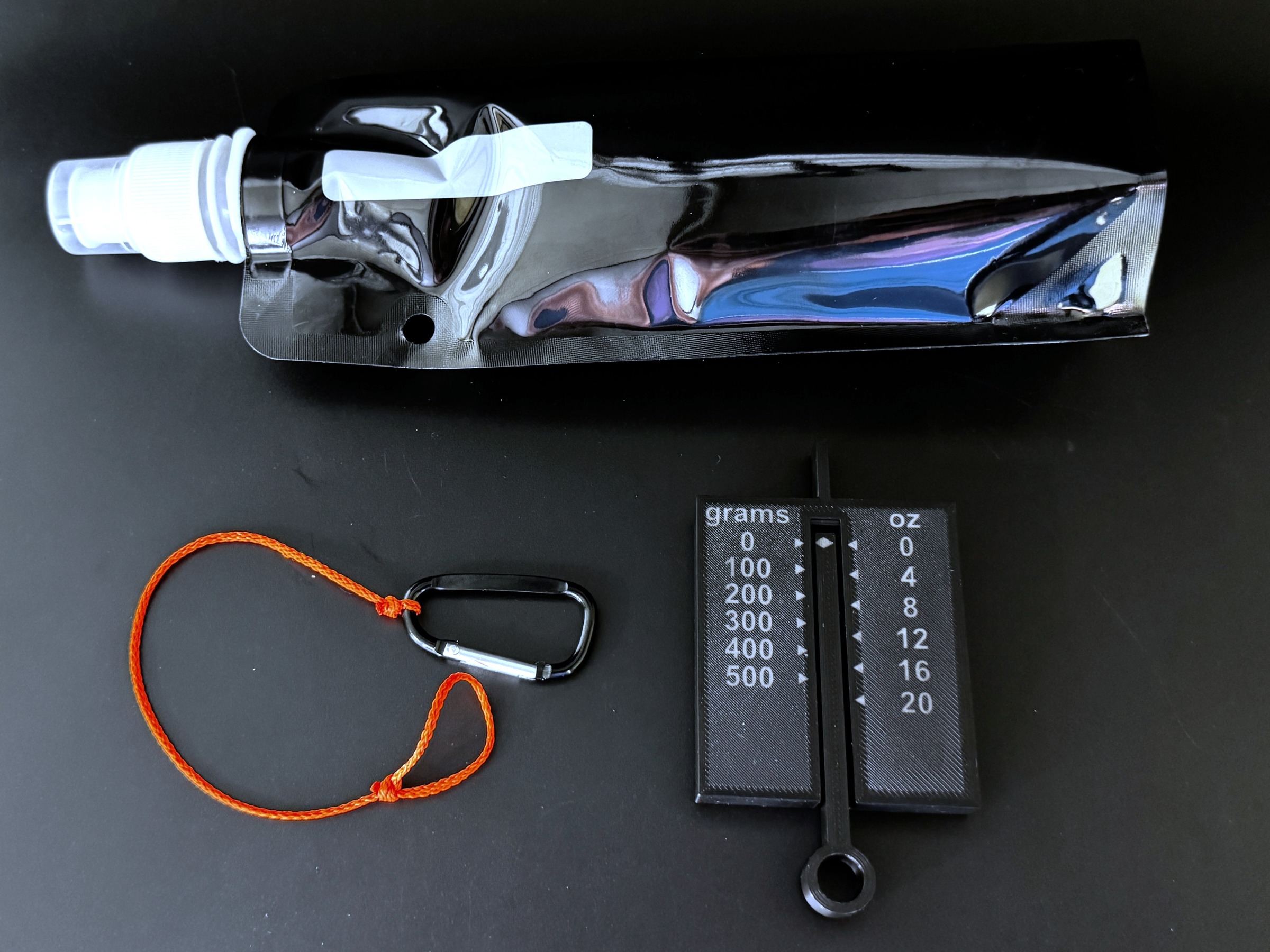 Weight Demo Kit contents: collapsible water bottle folded flat, weight gauge marked in grams and ounces, and orange tugging string with carabiner.