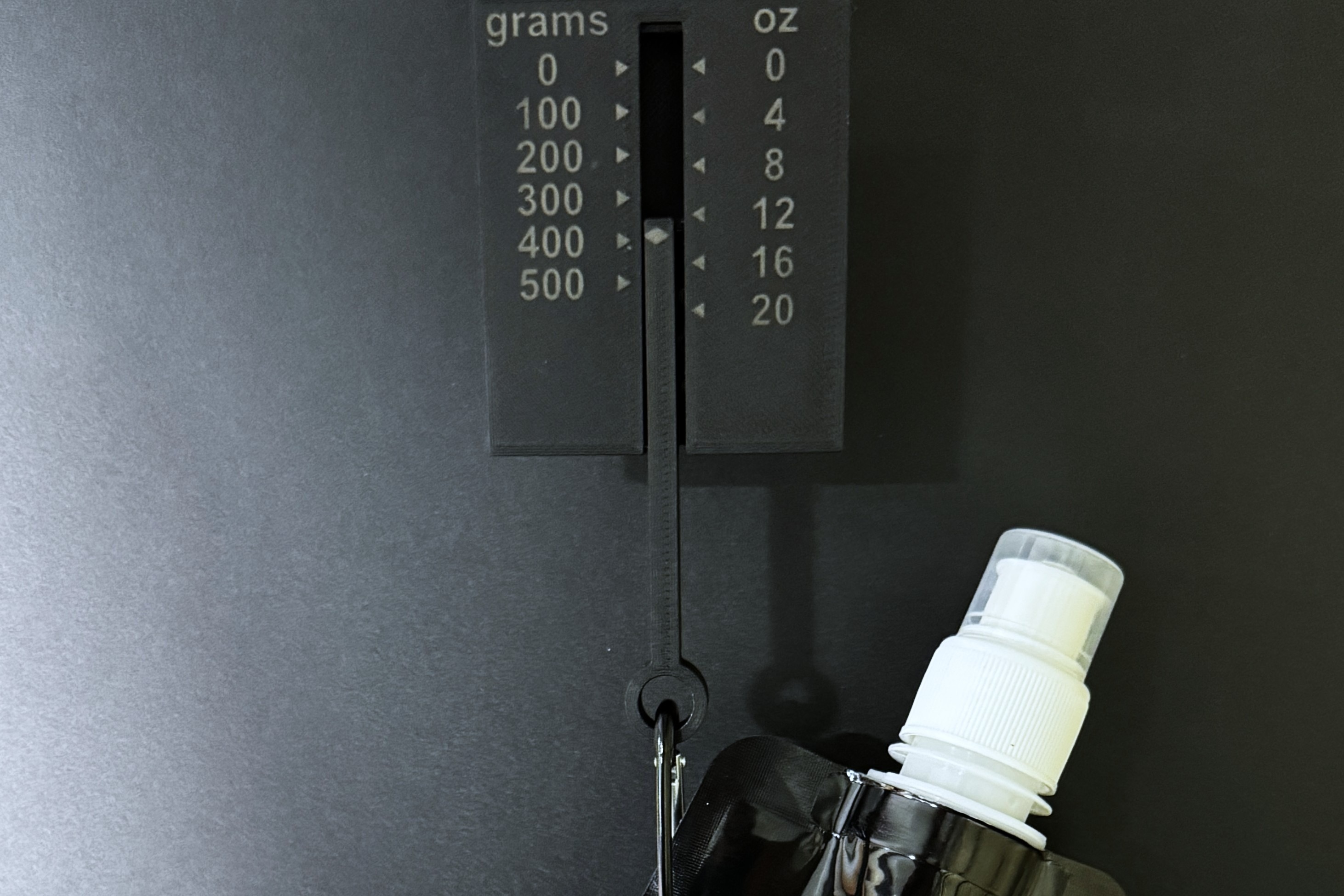 The collapsible water bottle clipped by its carabiner to the hanging tension gauge, which is marked in grams and ounces.