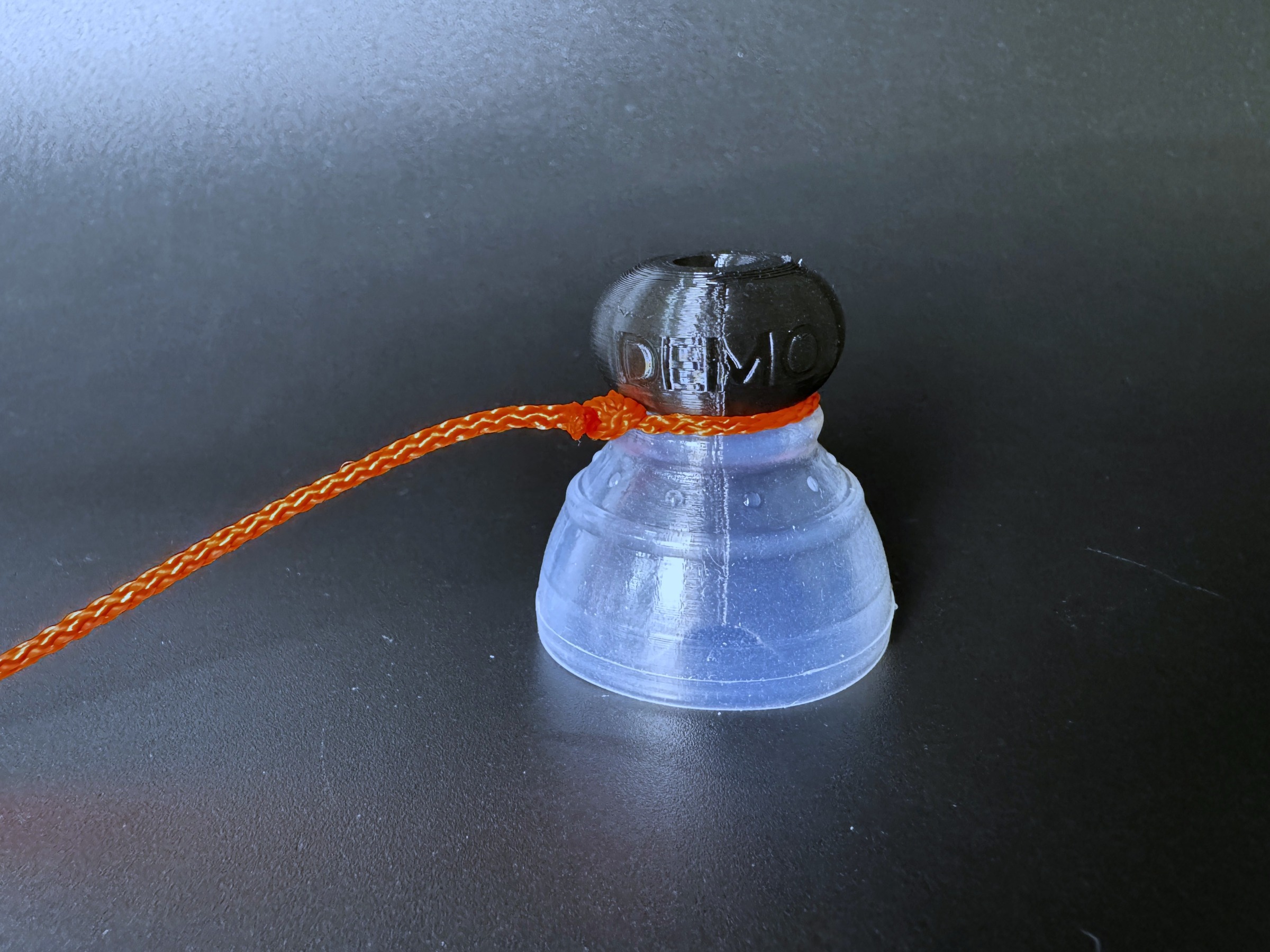 Orange tugging string looped around the knob end of a demo P-tainer fitted with a clear silicone gripper.
